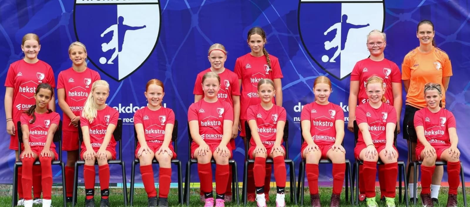 Meidenvoetbalteam in rode tenues voor een bedrukt spandoek van Dranghekdoek.nl